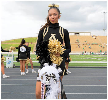 A person in a black and gold cheerleading uniform holds black and gold pom-poms and a decorated white mum while standing on a track near a football field with other cheerleaders and spectators in the background. Description generated by AI