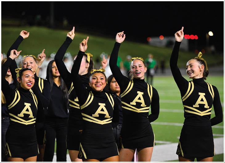 A group of adults in black and gold cheerleading uniforms with the letter A on the front are standing on a sports field at night with their arms raised and smiling. Description generated by AI