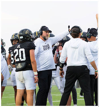 A person wearing a headset and a cap is gesturing with an outstretched arm while standing among people in football helmets. Description generated by AI