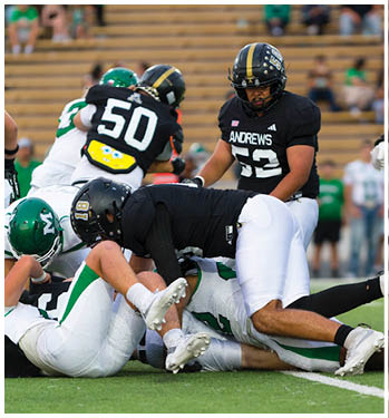 A person wearing a black football uniform with the word Andrews and the number 52 on the jersey is on a football field with blurred spectators in the background. Description generated by AI