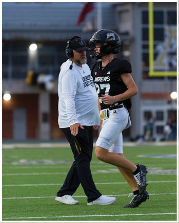 A person wearing a headset and white long-sleeve shirt is talking to a person in a black football uniform and helmet with the number 27 on it. Description generated by AI