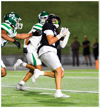 Two people in football uniforms are engaged in a play with one person in black catching the ball and the other in green and white attempting to defend. Description generated by AI