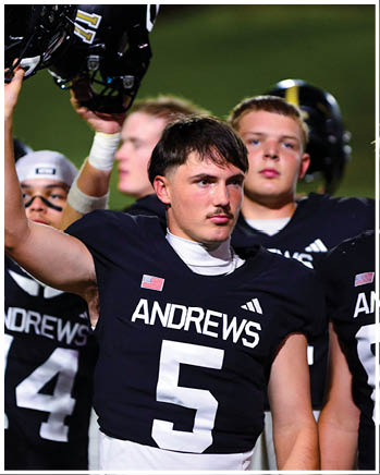 A person wearing a black football jersey with the name Andrews and number 5 is holding up a helmet while standing among other people in similar jerseys on a field. Description generated by AI