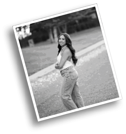 A person with long hair wearing a sleeveless top and jeans is standing outdoors on a path with trees in the background, smiling and looking over their shoulder. Description generated by AI