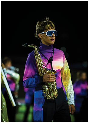 A person wearing a colorful gradient outfit and large reflective sunglasses is holding a saxophone on a dimly lit field with other people in the background. Description generated by AI