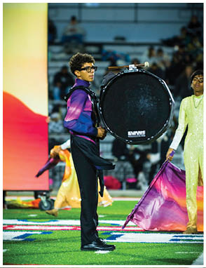 A person wearing a purple and blue uniform plays a large bass drum on a football field while another person in a yellow sequined outfit holds a purple and orange flag nearby. Description generated by AI