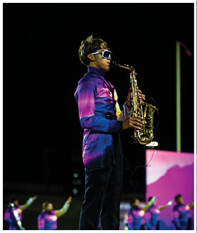 A person wearing a colorful uniform and sunglasses is playing a saxophone on a field with several other similarly dressed people in the background. Description generated by AI