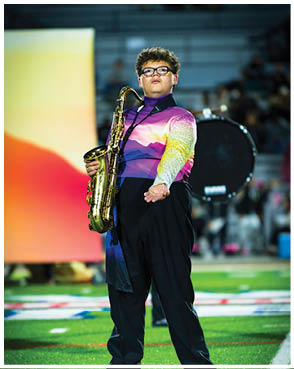 A person wearing a colorful uniform stands on a field holding a saxophone with one hand while extending the other hand forward. Description generated by AI