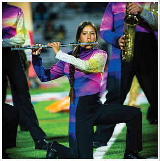 A person wearing a colorful uniform is playing a flute on a field with other musicians in the background. Description generated by AI