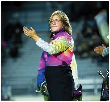 A person wearing glasses and a colorful, sparkly uniform is conducting or leading a band on a field at night. Description generated by AI