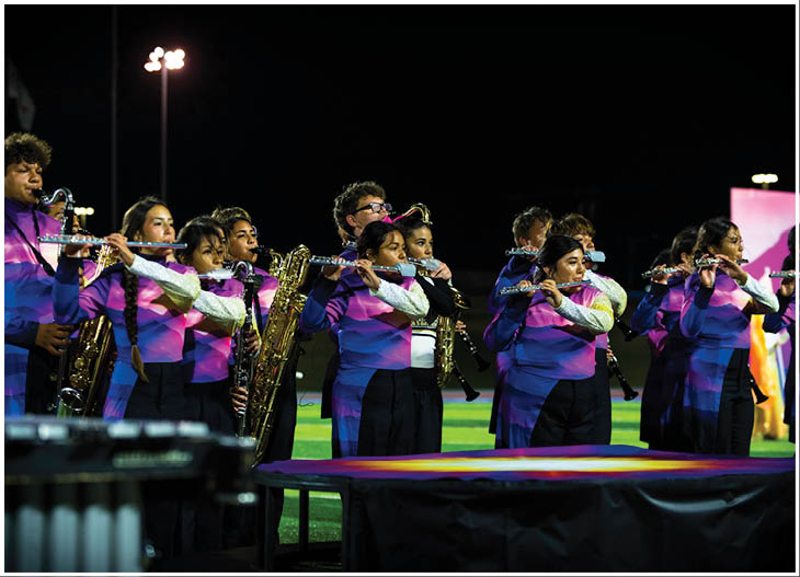 A group of people in matching purple and blue uniforms are playing wind instruments on a field at night. Description generated by AI