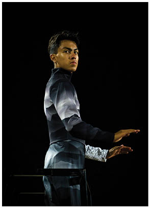 A person wearing a dark, patterned outfit with a high collar stands against a black background with arms extended forward in a poised stance. Description generated by AI