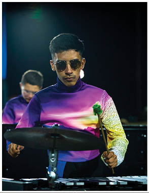 Four people wearing matching colorful gradient shirts are playing percussion instruments in a dimly lit setting, with one person in the foreground focused on striking a cymbal and xylophone. Description generated by AI