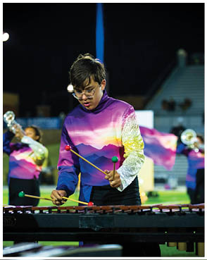 A person wearing a colorful purple and pink uniform is playing a marimba with mallets while other people in similar uniforms play brass instruments in the background on a field. Description generated by AI