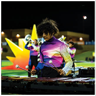 A person wearing a colorful uniform plays a marimba with mallets on a field while other people in similar uniforms play brass instruments in the background. Description generated by AI