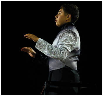 A person wearing a sequined silver and black uniform is conducting with raised hands against a dark background. Description generated by AI