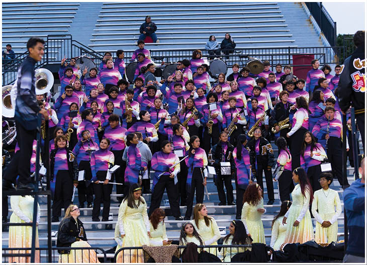 A large group of people wearing matching purple and pink gradient uniforms are seated in bleachers playing musical instruments while another group in light yellow costumes stands and sits in front of them. Description generated by AI