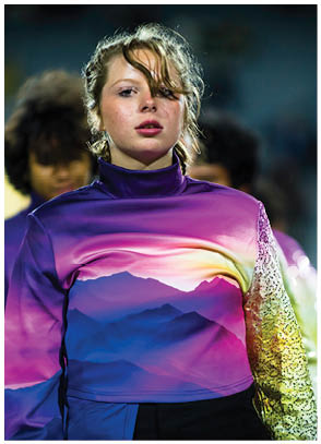 A person wearing a vibrant jacket with a mountain landscape design stands in front of others dressed in similar colorful attire, likely participating in a performance or event. Description generated by AI