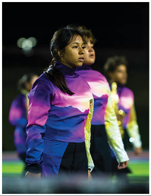 People wearing colorful gradient jackets are standing on a field, some holding musical instruments. Description generated by AI