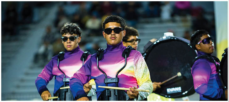 Three people wearing sunglasses and purple outfits are standing in front of a drum in a stadium setting. Description generated by AI