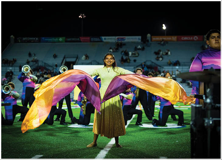 A person in a green and gold costume is performing with large flowing orange and purple flags on a football field while a marching band in purple and blue uniforms plays instruments in the background. Description generated by AI