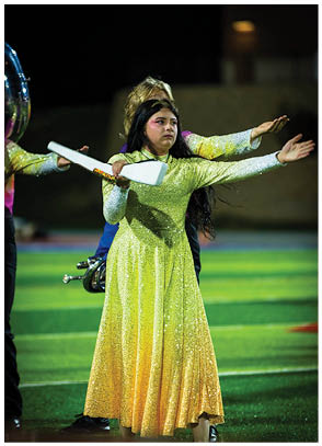 A person in a sparkling yellow and green gradient dress is performing a baton twirling routine on a field at night with other performers in the background. Description generated by AI