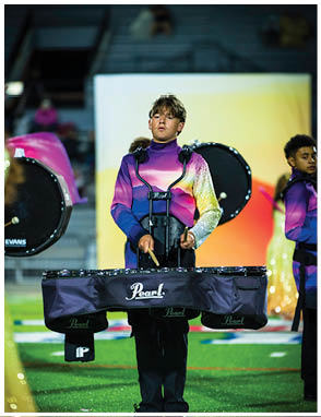 A person wearing a colorful purple and yellow uniform is playing a set of marching tenor drums on a field with other performers in the background. Description generated by AI