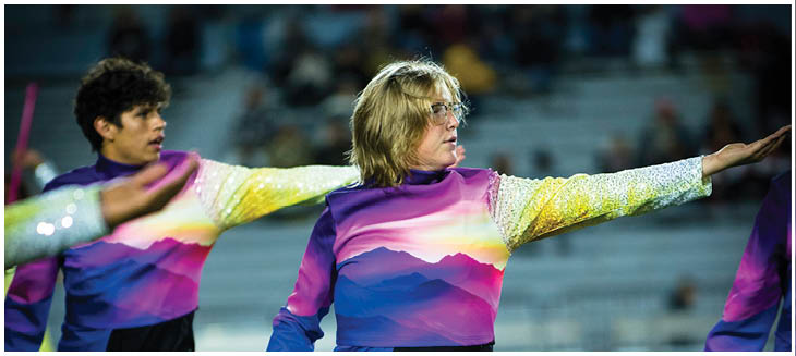 Two people wearing colorful, gradient long-sleeve outfits are extending their arms outward, appearing to perform a synchronized routine in a stadium setting. Description generated by AI