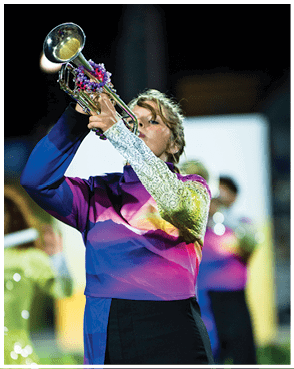 A person wearing a colorful uniform with a gradient pattern and lace sleeves is playing a trumpet during an outdoor performance. Description generated by AI