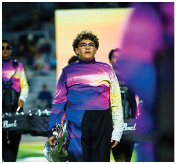 A person wearing a colorful gradient uniform and glasses is holding a brass instrument while walking on a field with other similarly dressed people in the background. Description generated by AI