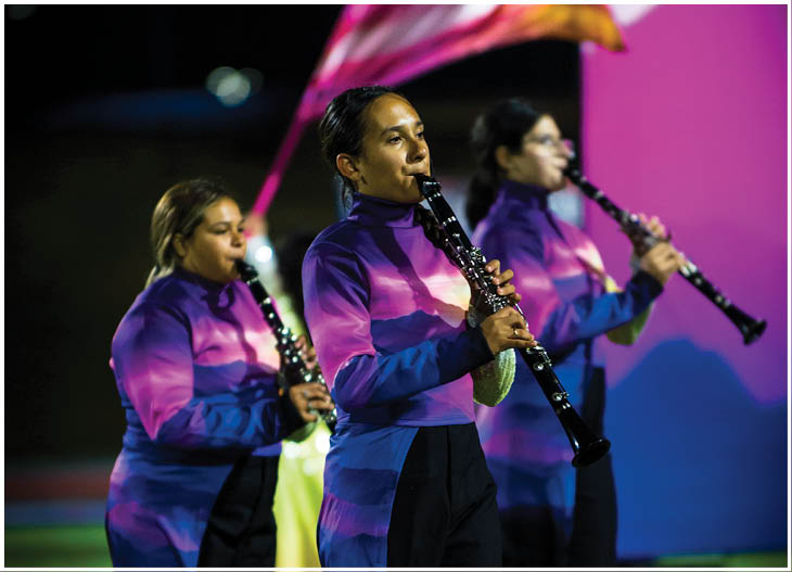 People in purple uniforms are playing clarinets with a blurred flag waving in the background against a dark sky. Description generated by AI