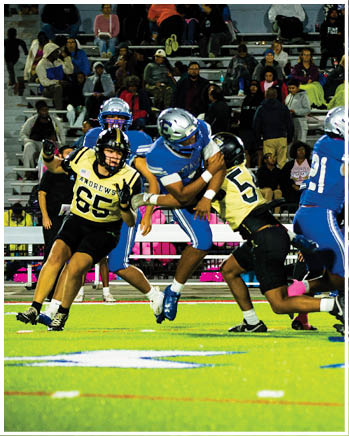 Players in blue and yellow football uniforms are engaged in a play on a brightly lit field with spectators watching from the bleachers. Description generated by AI