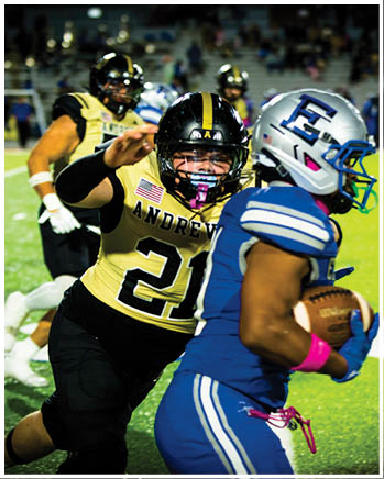 A person in a black and gold football uniform with the number 21 is reaching out toward another person in a blue and white football uniform holding a football on a field during a game. Description generated by AI