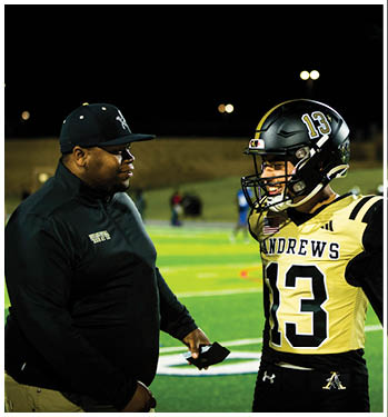 An adult wearing a black cap and jacket is talking to a person in a football uniform with the number 13 and the name Andrews on the jersey on a sports field at night. Description generated by AI