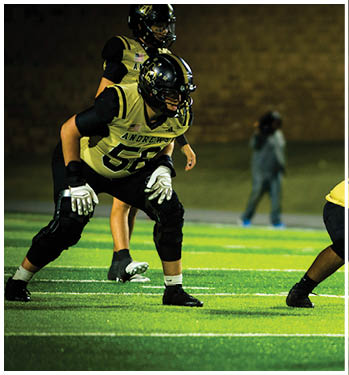 Three people in football uniforms and helmets are positioned on a field, appearing ready for a play during a nighttime game. Description generated by AI
