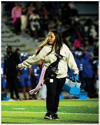 A person wearing glasses and a headset stands on a sports field with a pink jacket tied around their waist while a group of people in blue sports uniforms and coaches stand in the background near empty bleachers. Description generated by AI