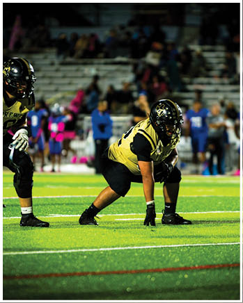 Several people in football uniforms are positioned on a field, appearing ready for a play during a game. Description generated by AI