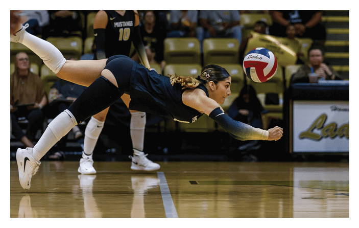 A person in athletic clothing is diving horizontally in mid-air to hit a volleyball on an indoor court with spectators in the background. Description generated by AI