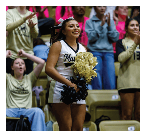 A person in a cheerleading uniform holding gold and black pom-poms is smiling while standing in front of a crowd of people who are clapping and cheering. Description generated by AI