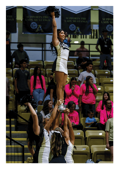 A person in a cheerleading uniform is smiling and raising one arm with a pom-pom in a gymnasium setting. Description generated by AI