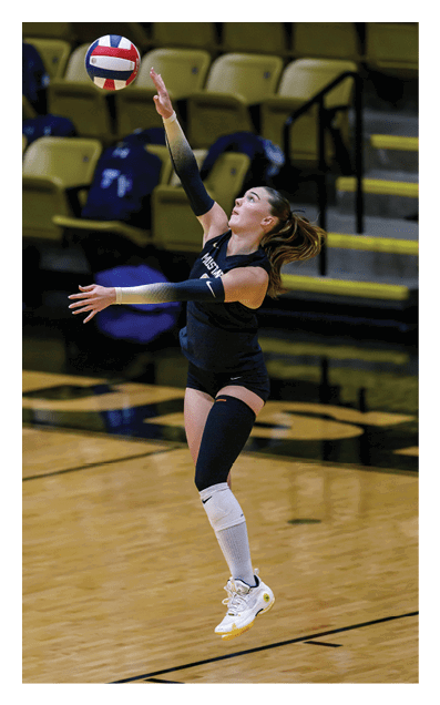 A person wearing a dark sports uniform and knee pads is jumping and reaching up to hit a volleyball in an indoor gymnasium. Description generated by AI