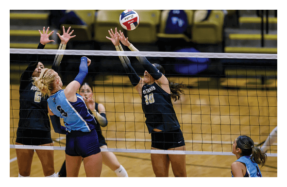People in athletic uniforms are actively playing volleyball indoors with two players jumping to block the ball at the net while another player prepares to hit it. Description generated by AI