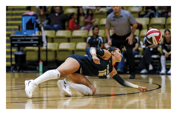 A person in athletic clothing is diving on a gym floor to hit a volleyball while others watch in the background. Description generated by AI