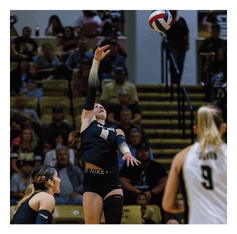 A person wearing a black sports uniform with the number 8 is reaching up to hit a volleyball in an indoor gymnasium with spectators in the background. Description generated by AI