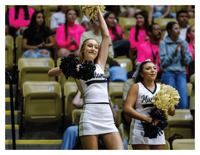 Two adults in cheerleading uniforms hold pom-poms and perform in front of seated spectators wearing bright pink shirts. Description generated by AI