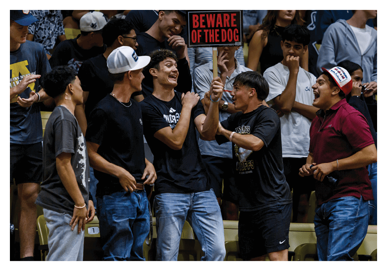 A group of people wearing casual clothing are enthusiastically interacting and holding a sign that says “BEWARE OF THE DOG" in a crowded indoor setting. Description generated by AI
