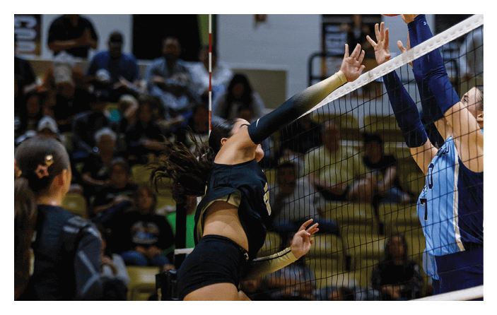 Two people in athletic clothing are jumping near a volleyball net with one person attempting to block or hit the volleyball while the other reaches to intercept it during a game in an indoor gymnasium. Description generated by AI
