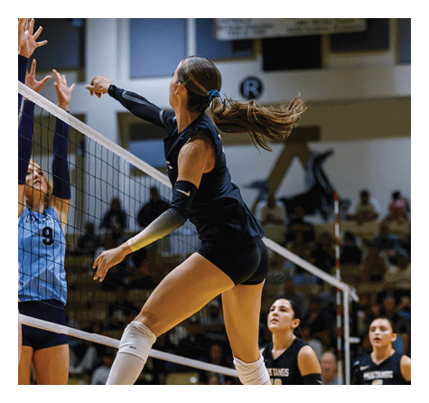 A person with a ponytail wearing a dark athletic outfit is jumping near a volleyball net, reaching out to hit a volleyball in an indoor gymnasium. Description generated by AI
