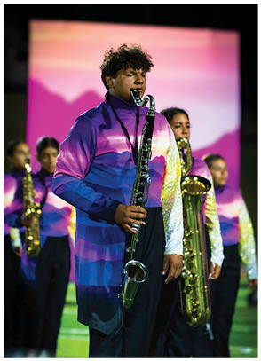 A group of people wearing colorful gradient uniforms are playing saxophones on a field with a blurred purple and pink background. Description generated by AI