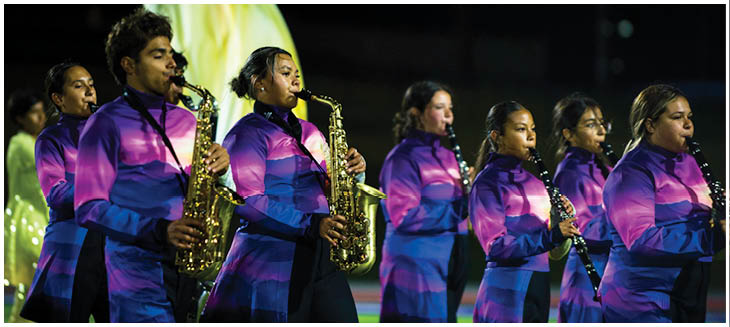 A group of people wearing matching purple and blue uniforms are playing saxophones in a line, likely performing at an outdoor event at night. Description generated by AI